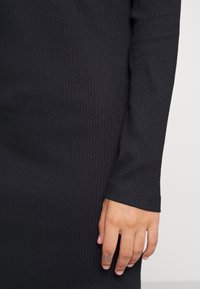 Zwart ribbeljurk met lange mouwen en een getailleerde pasvorm. Heeft een soepele textuur en een nette zoom, met een hand die op de stof rust.