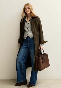 Young woman in long dark brown coat, gray knit cardigan, blue wide-leg jeans, holding woven brown leather handbag, standing against neutral wall.