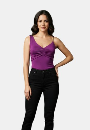 Donna con capelli scuri e mossi che indossa un top viola senza maniche con arricciature e pantaloni neri a vita alta, in piedi con una mano sul fianco.