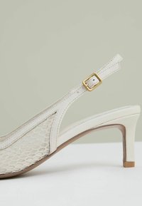 Slingback podpatky z bílé kůže s síťovinovou panelem, zdobené zlatou sponou a štíhlým, zužujícím se podpatkem. Texturovaný design zvyšuje vizuální přitažlivost.