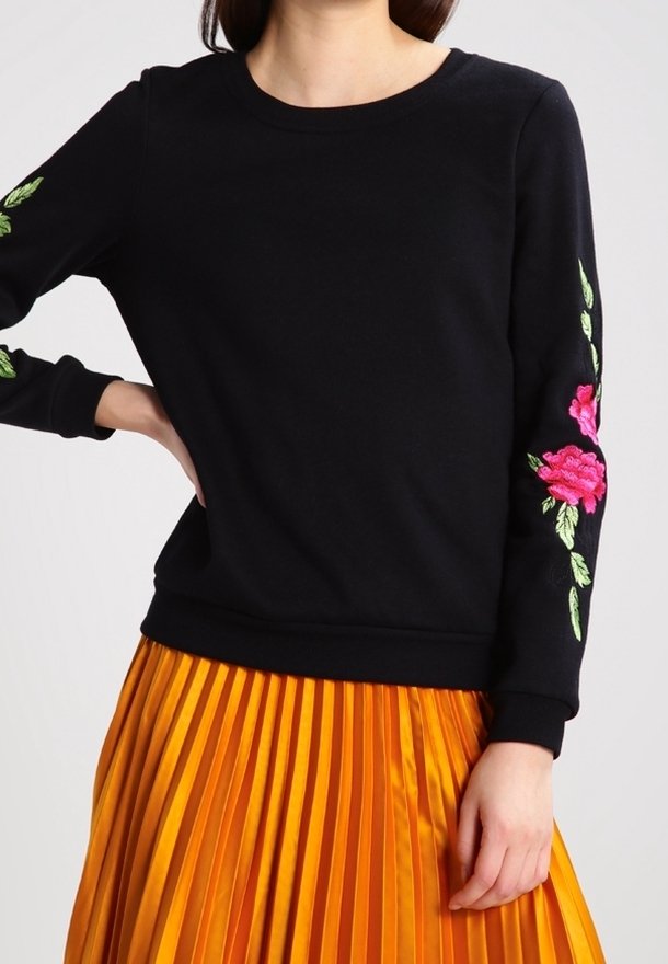 Femme portant un haut noir à manches longues avec une broderie florale rose sur les manches et une jupe plissée orange vif, debout avec une main sur la hanche.