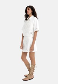 Biała, krótkowana bluzka z perforowanymi kwiatowymi detalami, połączona z białą spódnicą z podobnymi wycięciami. Beżowe sandały na sznurówki dopełniają stylizację.