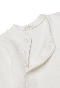 Camisa de algodón blanca con cuello redondeado, cierre de botones a presión y textura suave. Etiqueta de cuidado visible cerca del escote.