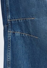 Μπλε ντένιμ ύφασμα με διπλές ραφές και δύο οριζόντιες, ελαφρώς ζαρωμένες πλαϊνές τσέπες σε ένδυμα.