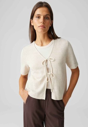 Donna con capelli castani lisci indossa un cardigan crema a maniche corte legato con due fiocchi sopra una maglietta bianca, abbinato a pantaloni marrone scuro, con le mani in tasca.