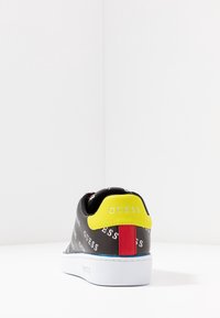 Baskets en cuir synthétique noir avec une semelle blanche. Fonctionnalités: accent de talon jaune, stripe vertical rouge et motif logo "GUESS" en blanc répété.