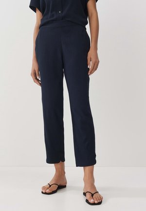 Frau trägt marineblaue, verkürzte, strukturierte Hose mit seitlichen Schlitzsäumen und schwarze Zehensandalen, steht vor einfarbigem Hintergrund.