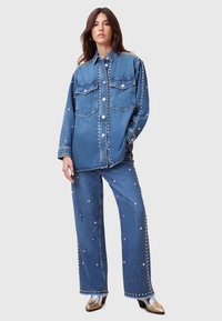 Conjunto de chaqueta y pantalones de mezclilla con tachuelas plateadas, dos bolsillos en el pecho y cierre frontal de botones. Corte holgado, color azul claro y textura suave.