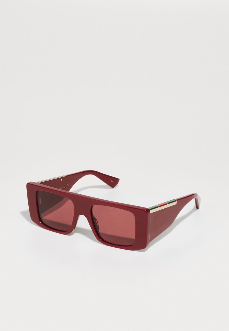 Ochelari de soare supradimensionați, roșii, cu rame dreptunghiulare, lentile colorate și accente de hardware aurii. Bratele prezintă un detaliu cu dungi multicolore.
