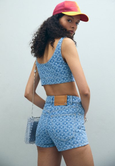 Top scurt din denim albastru cu model, asociat cu şorturi înalte asortate. Prezintă o etichetă din piele de culoare bej şi o geantă transparentă cu curea din lanț.