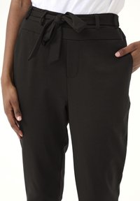 Zwarte broek van gladde stof, met een strikceintuurdetail in de taille en zijzakken. De pasvorm is ontspannen en op maat gemaakt.