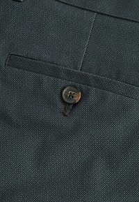 Tissu texturé foncé avec une petite poche et un bouton à quatre trous. Le bouton est noir avec un léger éclat, présentant un fil contrastant.