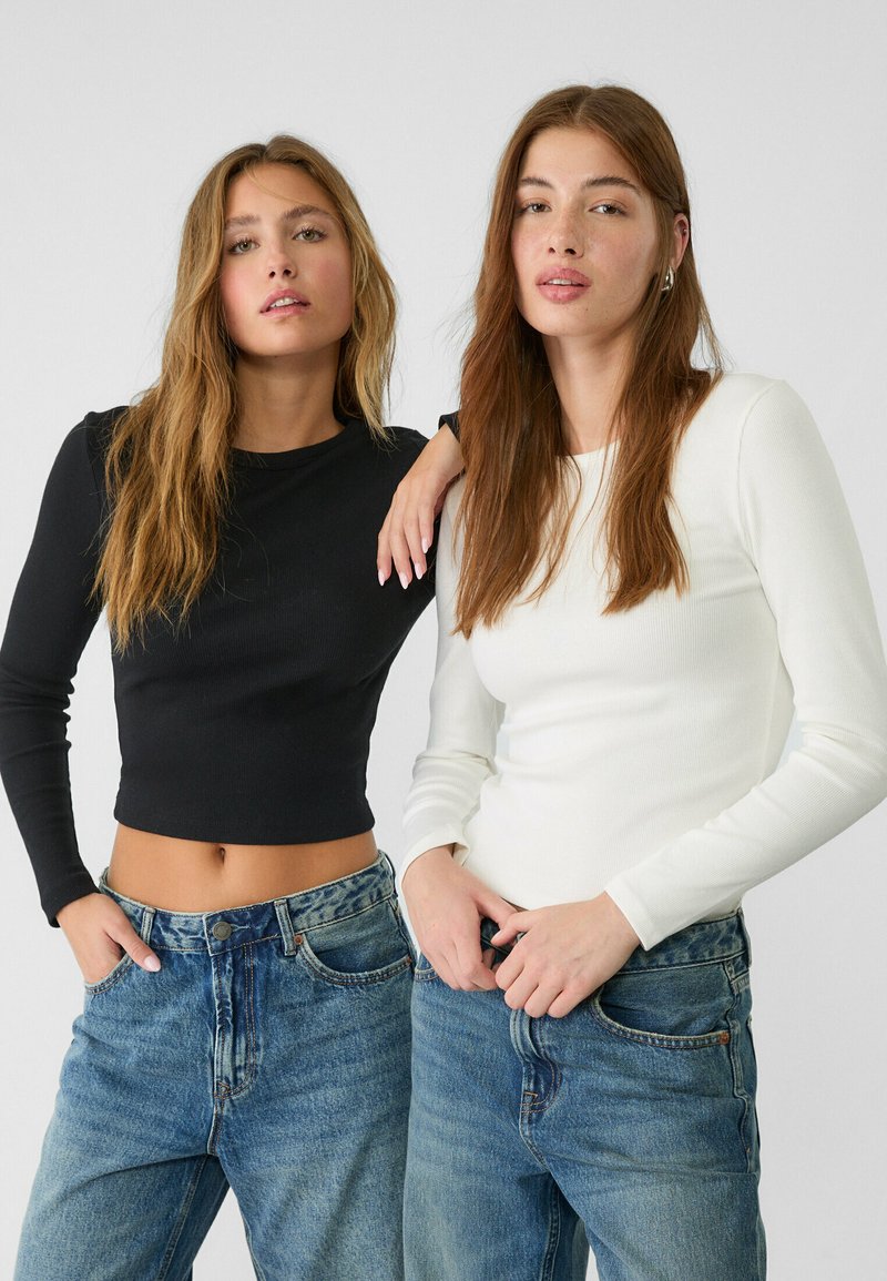Zwei Models tragen figurbetonte Langarmoberteile: eines in Schwarz und eines in Weiß, kombiniert mit hochgeschnittenen blauen Jeans. Glatte Stofftextur.