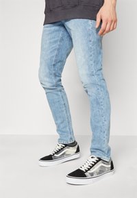 Light blue denim jeans with a slim fit, featuring subtle whiskering and a smooth texture, paired with black and white sneakers with a transparent overlay.