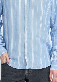 Chemise à boutons à rayures bleu clair avec des rayures verticales blanches, tissu texturé, et une étiquette sur l'ourlet inférieur, associée à un jean foncé.