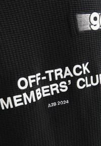 Sort tekstureret stof med hvid broderi, der læser "OFF-TRACK MEMBERS' CLUB A2B 2024," med et logoelement ovenfor.