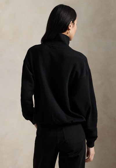 Woman with long dark hair wearing black turtleneck sweater and black pants, standing with her back to the camera against neutral background.