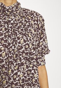 Bordeauxfarbige Bluse mit floralem Muster in Creme und Gelb, mit gerafftem Ausschnitt und kurzen Ärmeln. Glatte Stoffqualität.