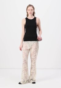 Haut sans manches noir associé à un pantalon évasé en dentelle crème avec un motif floral. Le mannequin tient un petit sac et se tient devant un fond neutre.
