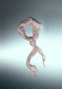 Světle růžový průsvitný šátek, uvázaný smyčkou s uzlem uprostřed; měkká textura a splývavé okraje s mírným mačkáním, na kovovém pozadí.