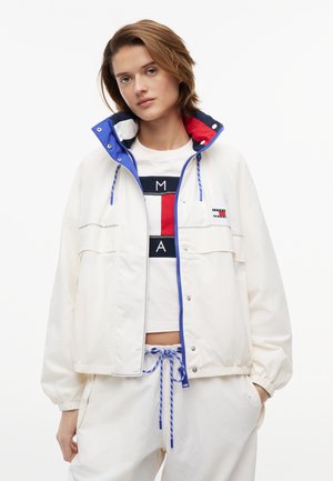 Kvinde iført hvid jakke med blå og røde kravedetaljer, hvide bukser med blå snor og en hvid t-shirt med tryk.