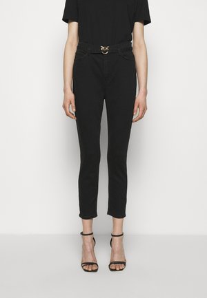 Persona con pantalones pitillo negros recortados, cinturón negro y sandalias negras de tacón con tiras, de pie frente a un fondo liso.