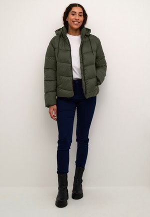 Chaqueta acolchada verde oliva con capucha y cremallera, con un diseño acolchado. Llevada sobre una camisa blanca y combinada con jeans azul oscuro y botas negras.