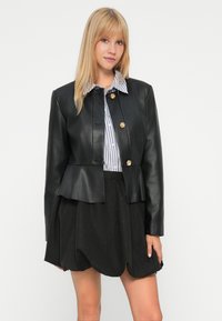Femme portant une veste en simili cuir noir avec des boutons dorés, une chemise rayée et une jupe noire texturée sur un fond blanc uni.