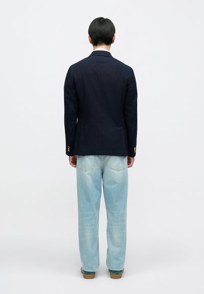 Granatowa marynarka z ozdobnymi guzikami, zestawiona z jasnoniebieskimi, luźnymi dżinsami oraz zielonymi butami. Minimalistyczny design z czystymi liniami.