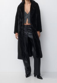 Cappotto in pelliccia sintetica nera con grande collo, chiusura con bottoni e vestibilità oversize, indossato sopra un top corto brillante e pantaloni in ecopelle a vita alta.