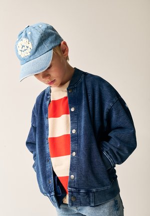 Jeans bomberjakke i mørkeblå, med knapper og ribbede manchetter. Underneath en beige skjorte med røde striber. Blå denimkasket med broderet logo.