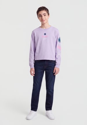 Adolescent debout portant une chemise à manches longues violet clair avec des motifs, un jean foncé et des baskets blanches sur un fond uni.