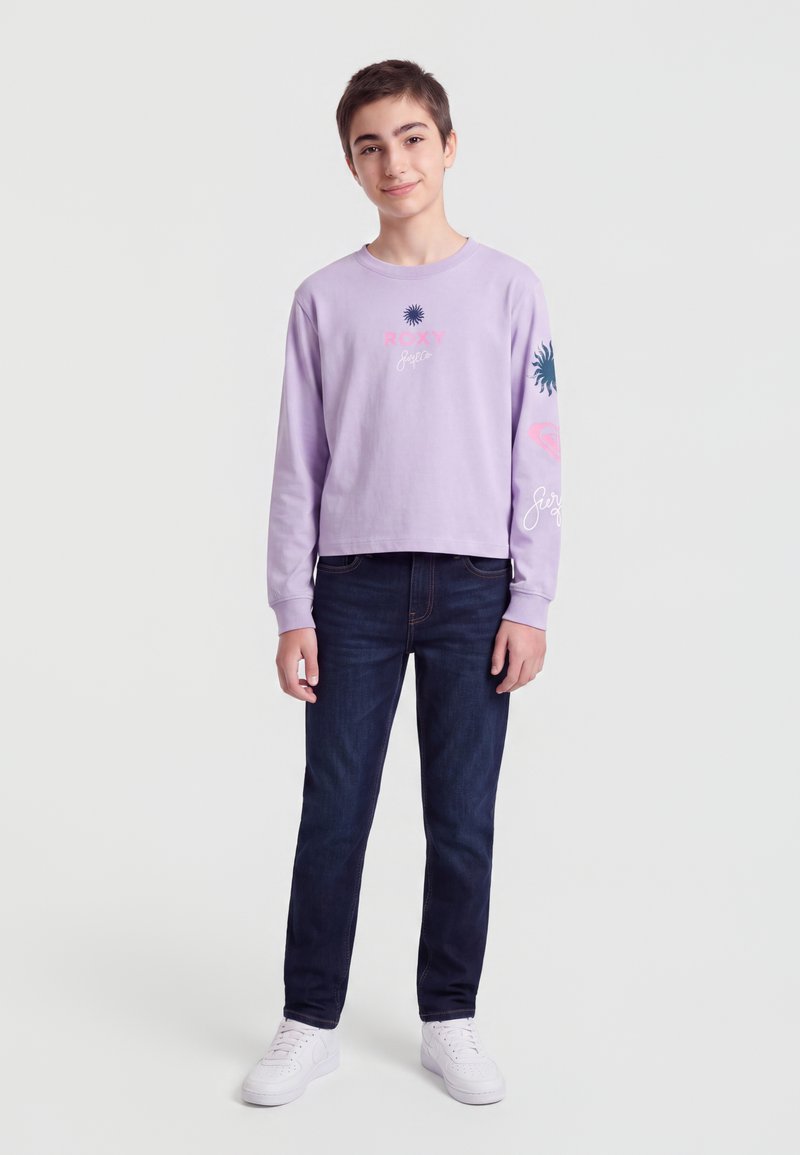 Adolescent debout portant une chemise à manches longues violet clair avec des motifs, un jean foncé et des baskets blanches sur un fond uni.