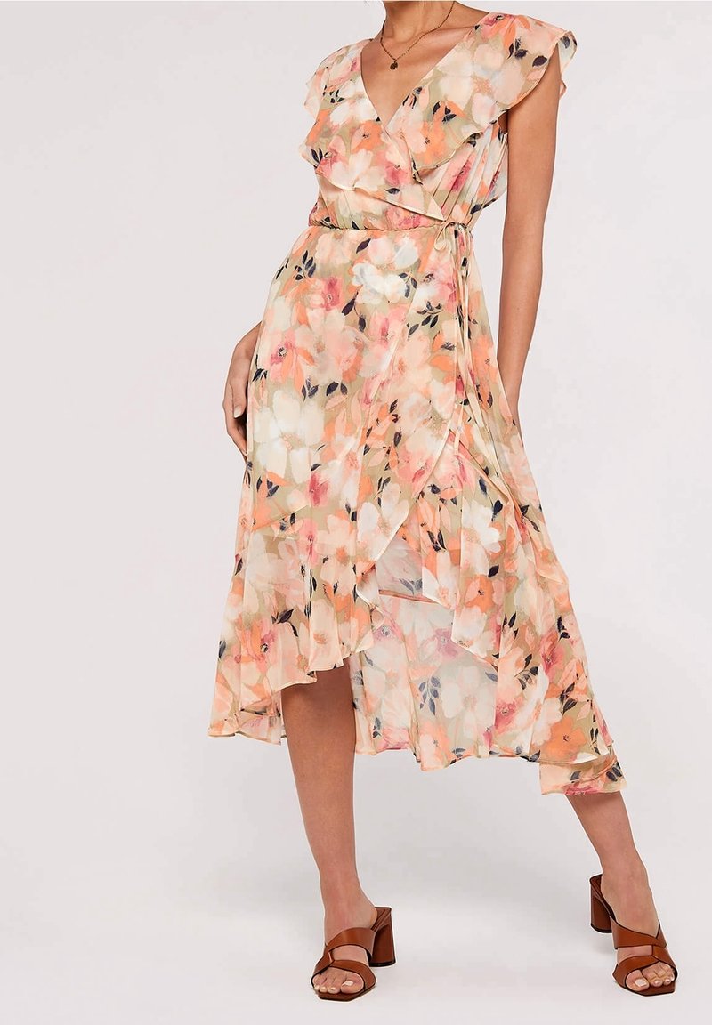 Femme portant une robe portefeuille fleurie sans manches avec des volants et des sandales à talons blocs couleur cuir clair sur un fond uni.