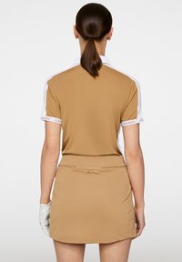 Ruskea golfmekko lyhyillä hihoilla, valkoisilla yksityiskohdilla ja brändätyillä hihansuilla. Mekossa on istuva vyötärö ja sileä tekstuuri.