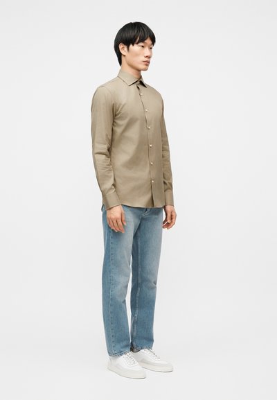 Νέος άνδρας που στέκεται φορώντας ένα μπεζ πουκάμισο με κουμπιά, ανοιχτό μπλε τζιν και λευκά αθλητικά παπούτσια μπροστά σε λευκό απλό φόντο.