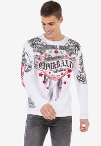 Langärmliges weißes T-Shirt mit schwarzen und roten Grafiken, darunter ein Wappenlogo und Sterne, mit einem lässigen Schnitt und rundem Ausschnitt.