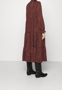 Maxi šaty s červeným květinovým vzorem, dlouhými rukávy, volánkovým lemem a vysokým výstřihem. V kombinaci s černými kovbojskými botami s precizním prošíváním.