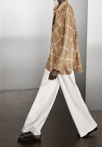 Bruin geruit overhemd met lange mouwen en knopen, gecombineerd met witte wijde broek en zwarte sandalen, tentoongesteld tegen een neutrale achtergrond.