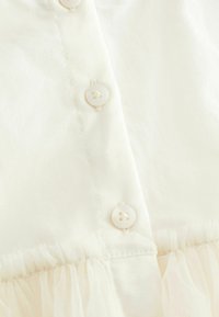 Blouse en coton blanc avec une rangée de cinq boutons ronds et transparents et un ourlet en couches avec volants. Texture lisse avec une légère brillance.