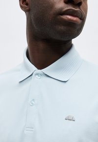 Gros plan d'un homme portant un polo bleu clair texturé avec un petit logo gris brodé sur la poitrine.