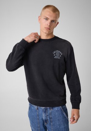 Jeune homme aux cheveux blonds courts portant un sweat-shirt noir avec le logo California Athletics et un jean bleu, debout devant un fond gris uni.