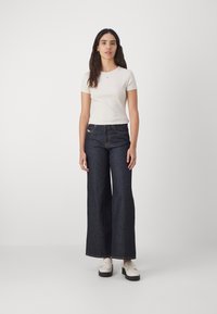Hvit kortermet t-skjorte kombinert med mørkeblå vide denimjeans og hvite loafers. Stoffet ser glatt ut med minimal dekor.