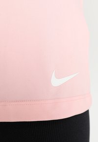 Parte superior deportiva Nike en rosa claro, con una textura suave y un logo Swoosh blanco cerca del dobladillo, combinada con pantalones negros.