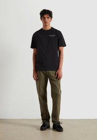 Czarna bawełniana koszulka z krótkimi rękawami i białym logo; zestawiona z oliwkowo-zielonymi spodniami i czarnymi butami o teksturowanej podeszwie.