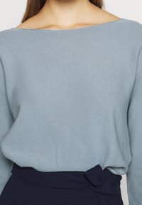 Světle modrý pletený svetr se širokým výstřihem, dlouhými rukávy a uvolněným střihem, kombinovaný s tmavě modrou sukní s vysokým pasem a mašlí.