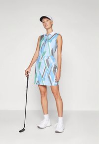 Nő kék, zöld és szürke mintás ujjatlan golfdresszben és fehér sapkában, golfütőt tartva, sima háttér előtt áll.