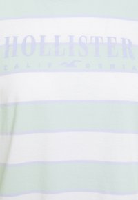 Pruhované tričko s jemně zelenými a bílými horizontálními pruhy, s velkým logem "HOLLISTER" v měkké levandulové barvě přes hruď.
