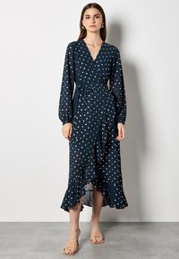 Vestito avvolgente blu navy con pois dorati, maniche lunghe, orlo a balze e vita con cinturino. Realizzato in tessuto leggero con una texture morbida.