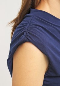 Granatowa bluzka z miękkiego materiału, z podkreślonymi bufiastymi rękawami i gładką fakturą. Zbliżenie uwypukla okolice ramion oraz szczegóły rękawów.